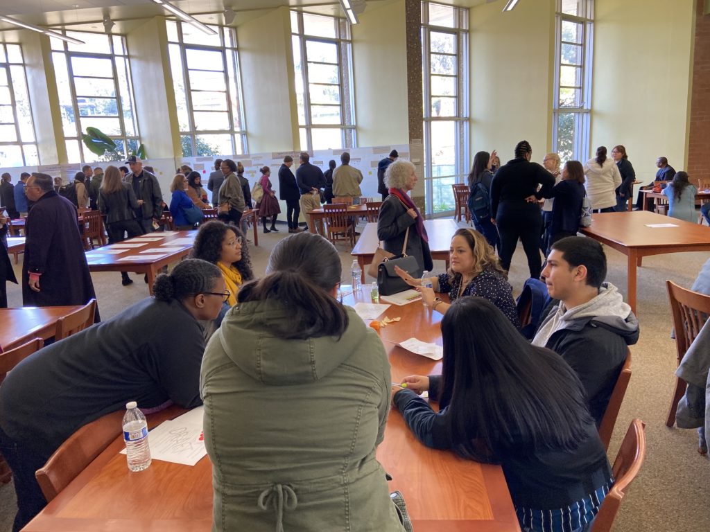 People at all college day, talking in groups and pairs in library.  Back of room has white paper with activity